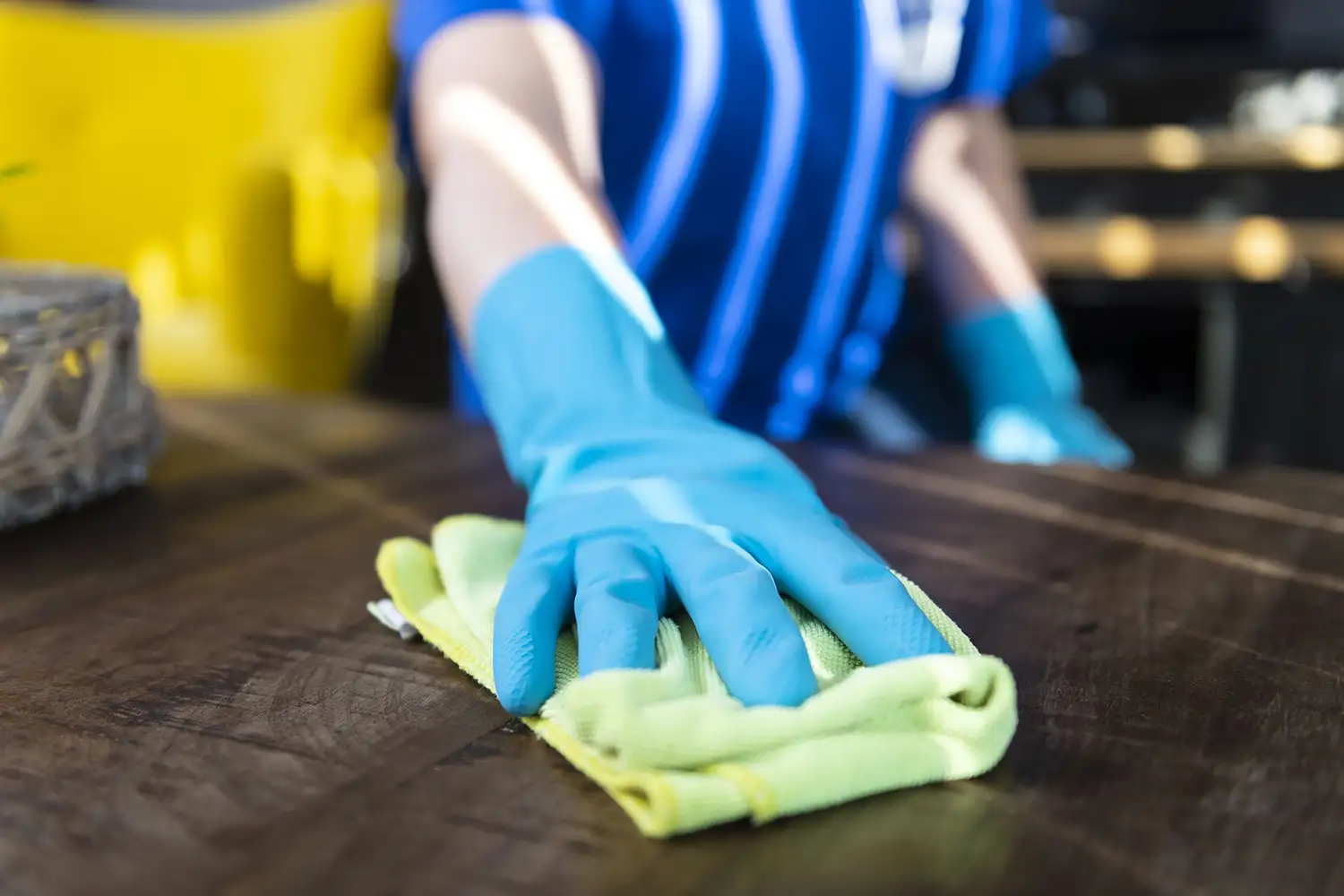 Une main gantée de bleu nettoie une table en bois avec un chiffon jaune, sur fond de chaise jaune floue. Meta description : Découvrez nos services de nettoyage professionnels pour un intérieur impeccable et soigné.